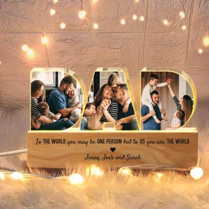 K&ouml;nnte beinhalten: Ein Holz-Bilderrahmen in Form des Wortes "DAD" mit einem Licht im Inneren. Der Rahmen enth&auml;lt drei Fotos einer Familie mit einem Vater und Kindern. Der Text "To the world you may be one person but to us you are the world" ist auf dem Rahmen gedruckt. Die Namen "Jenny, Josh und Sarah" sind unter dem Text gedruckt.