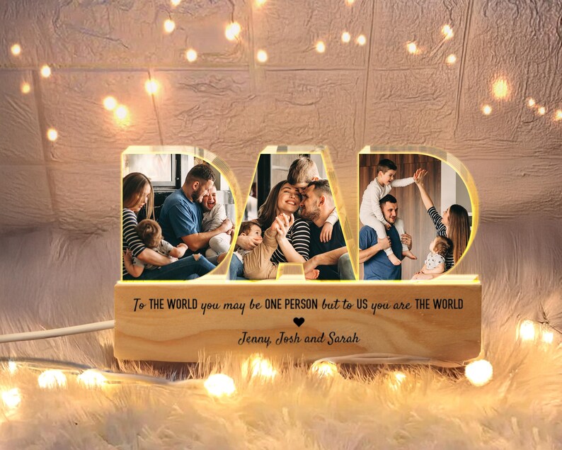 K&ouml;nnte beinhalten: Ein Holz-Bilderrahmen in Form des Wortes "DAD" mit einem Licht im Inneren. Der Rahmen enth&auml;lt drei Fotos einer Familie mit einem Vater und Kindern. Der Text "To the world you may be one person but to us you are the world" ist auf dem Rahmen gedruckt. Die Namen "Jenny, Josh und Sarah" sind unter dem Text gedruckt.