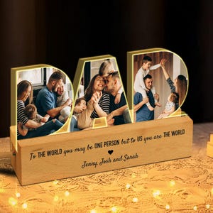 K&ouml;nnte beinhalten: Ein Holz-Bilderrahmen mit einem leuchtenden "DAD"-Design. Der Rahmen hat vier separate Fotof&auml;cher, die jeweils ein anderes Familienfoto zeigen. Die Basis des Rahmens tr&auml;gt den Text "To the world you may be one person but to us you are the world" und "Jenny, Josh and Sarah".