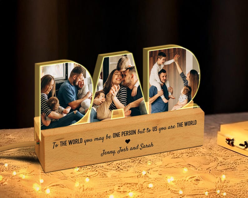 K&ouml;nnte beinhalten: Ein Holz-Bilderrahmen mit einem leuchtenden "DAD"-Design. Der Rahmen hat vier separate Fotof&auml;cher, die jeweils ein anderes Familienfoto zeigen. Die Basis des Rahmens tr&auml;gt den Text "To the world you may be one person but to us you are the world" und "Jenny, Josh and Sarah".