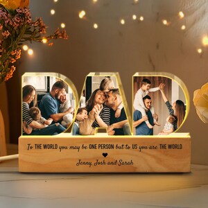 K&ouml;nnte beinhalten: Ein beleuchtetes Holzschild mit dem Wort "DAD" in gro&szlig;en Buchstaben. Die Buchstaben werden mit warmwei&szlig;en Lichtern beleuchtet. Jeder Buchstabe enth&auml;lt ein Foto einer Familie. Das Schild tr&auml;gt die Botschaft "To the world you may be one person but to us you are the world." Die Botschaft wird von den Namen "Jenny, Josh und Sarah" gefolgt.