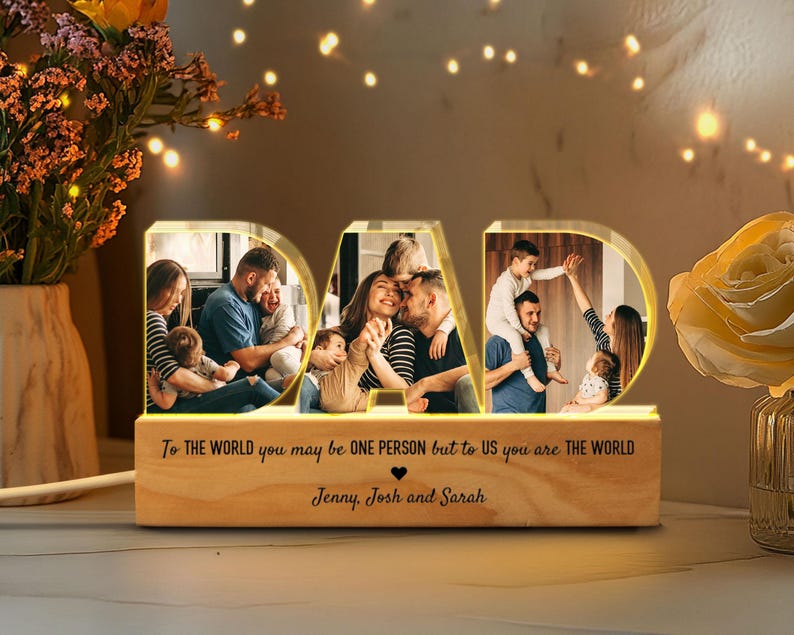 K&ouml;nnte beinhalten: Ein beleuchtetes Holzschild mit dem Wort "DAD" in gro&szlig;en Buchstaben. Die Buchstaben werden mit warmwei&szlig;en Lichtern beleuchtet. Jeder Buchstabe enth&auml;lt ein Foto einer Familie. Das Schild tr&auml;gt die Botschaft "To the world you may be one person but to us you are the world." Die Botschaft wird von den Namen "Jenny, Josh und Sarah" gefolgt.