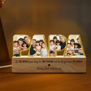 K&ouml;nnte beinhalten: Ein Holz-Bilderrahmen mit einem leuchtenden "PAPA"-Design. Der Rahmen zeigt ein Familienfoto und eine personalisierte Nachricht: "F&uuml;r die Welt bist du vielleicht nur eine Person, aber f&uuml;r uns bist du die Welt." Die Nachricht wird von den Namen "Jenny, Josh und Sarah" gefolgt.