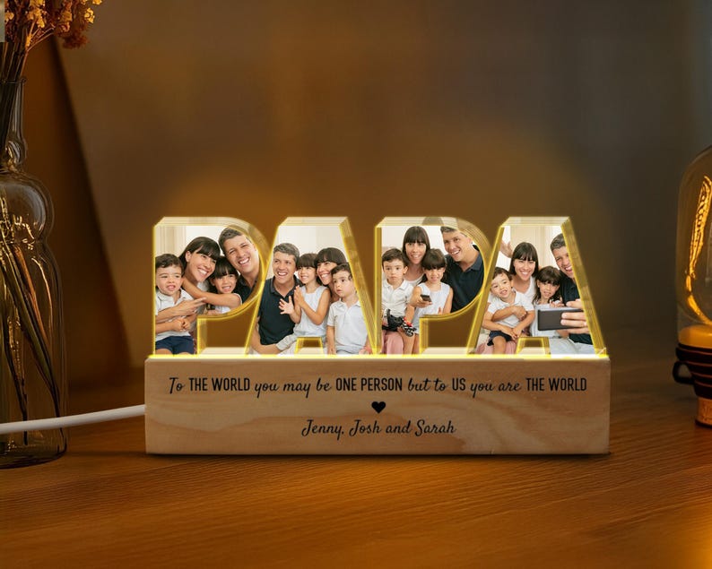 K&ouml;nnte beinhalten: Ein Holz-Bilderrahmen mit einem leuchtenden "PAPA"-Design. Der Rahmen zeigt ein Familienfoto und eine personalisierte Nachricht: "F&uuml;r die Welt bist du vielleicht nur eine Person, aber f&uuml;r uns bist du die Welt." Die Nachricht wird von den Namen "Jenny, Josh und Sarah" gefolgt.