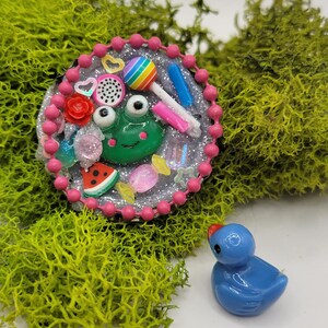 May include: A round, pink-rimmed resin charm featuring a green frog face, rainbow lollipop, watermelon slice, and other colourful candy decorations. The charm is resting on a bed of green moss.