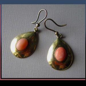 May include: A pair of teardrop-shaped earrings. Each earring features a gold-toned metal frame with a smooth, polished finish. A coral-colored oval stone is set in the center of each earring. The earrings have a simple hook closure.