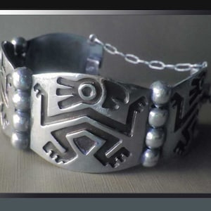 May include: A silver-toned bracelet with an Aztec-inspired design. The bracelet features geometric patterns and a chain closure. The design includes stylized figures and shapes, with a polished finish. The bracelet is a piece of jewelry.