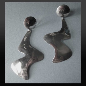 May include: A pair of silver earrings. Each earring has a small, round stud and a larger, abstract, wave-like shape. The earrings have a polished, reflective surface. They are displayed against a grey background.