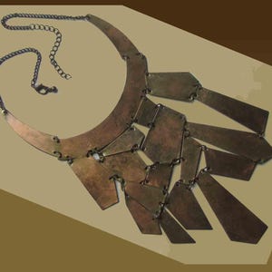 May include: A bronze-toned statement necklace with a curved collar and a series of rectangular and trapezoidal pendants. The necklace has a chain closure and is displayed against a neutral background. The metal has a textured appearance.