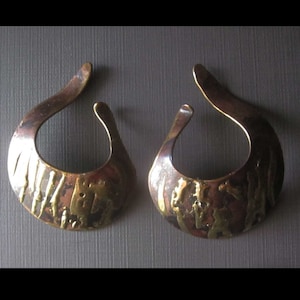 May include: A pair of bronze-colored earrings with a teardrop shape. The earrings have a textured surface with a mix of bronze and gold tones. The design is open at the top and bottom.