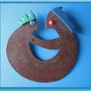 Könnte beinhalten: Eine skurrile, abstrakte Metallskulptur mit zwei stilisierten Figuren, die sich umarmen. Eine Figur hat eine grüne Krone und ein blaues Auge, die andere hat blaue Haare und ein rotes Auge. Die Skulptur ist rostbraun und vor blauem Hintergrund.