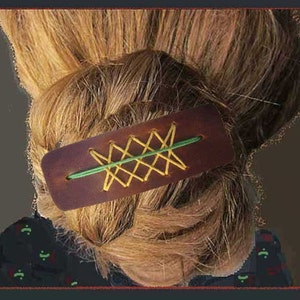 May include: A brown leather hair barrette with yellow and green stitching is shown in the image. The barrette is rectangular and is holding up a bun of light brown hair. The stitching creates a decorative pattern.
