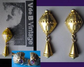 Puede incluir: Pendientes vintage dorados con diseño geométrico. Cada pendiente presenta una parte superior redondeada con una pequeña piedra púrpura, una sección central con recortes rectangulares y un colgante en forma de lágrima. Cierre de poste.