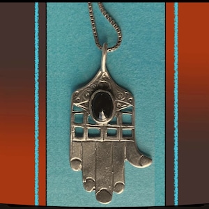 May include: Silver-toned Hamsa hand pendant necklace with an oval black stone. The pendant features intricate cut-out designs and is suspended from a delicate chain. The background is a mix of teal, orange, and brown.