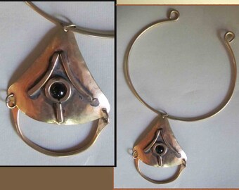 Puede incluir: Un collar de latón hecho a mano con un colgante de forma triangular, una piedra negra y un diseño circular. El collar tiene un collar abierto de color dorado. El colgante está sujeto al collar con una pequeña cadena.