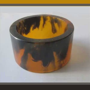 May include: A cylindrical ring with a marbled pattern of black, brown, and amber. The ring is made of a solid material and has a smooth, polished surface. The interior of the ring is a bright amber color.
