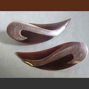 May include: Two vintage leaf-shaped earrings. Each earring features a dark brown, curved base with a silver-colored, swirling accent. The earrings have a polished finish and are set against a light gray background.