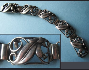 Puede incluir: Una pulsera plateada con diseño Art Nouveau. La pulsera presenta una serie de eslabones interconectados, cada uno adornado con motivos florales y de hojas estilizados. El cierre es visible en el primer plano, mostrando los intrincados detalles del trabajo en metal.