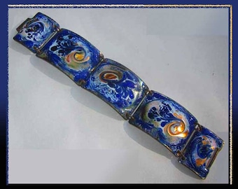 Puede incluir: Un brazalete azul y dorado con un diseño abstracto. El brazalete está hecho de varios eslabones rectangulares, cada uno con un patrón arremolinado en tonos de azul, blanco y dorado. El metal tiene una apariencia oscura y envejecida.