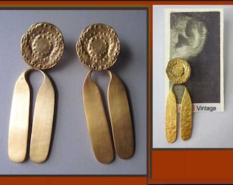Puede incluir: Pendientes dorados con una parte superior circular texturizada y una parte inferior alargada y curvada. Los pendientes tienen un aspecto de metal martillado. Una versión más pequeña de los pendientes se muestra junto a una imagen vintage con la palabra "Vintage".