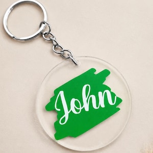 May include: A circular clear acrylic keyring featuring a vibrant green brushstroke graphic in the centre. The name John is printed in white cursive script over the green shape. The keyring includes a silver metal split ring and a short chain.