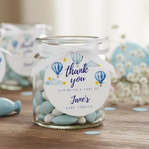 May include: A clear glass jar filled with blue and white candies, tied with a white ribbon and a circular tag. The tag features hot air balloons, stars, and the text "thank you for being a part of Jane's baby shower."