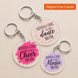 Peut inclure: Trois porte-clés circulaires en acrylique transparent avec des anneaux en métal argenté. Chaque pièce présente un fond en forme de coup de pinceau rose, rose vif ou violet clair, avec le texte noir KINDA CHIC TO BE A DANCE MOM, CHEER MOM ou MUSIC MOM.