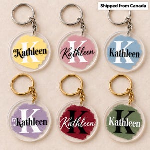 Peut inclure: Six porte-clés circulaires en acrylique avec des anneaux en métal argenté ou doré. Chaque pièce présente un fond peint en forme de coup de pinceau jaune, rose, bleu, violet, rouge ou vert, avec une lettre K et le nom Kathleen en écriture.