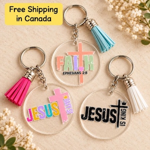 May include: Three circular clear acrylic keychains with metal rings and colourful tassels. One features a peach cross and the text FAITH EPHESIANS 2:8, another has a pink cross and the text JESUS, and the third has a black cross and JESUS IS KING.