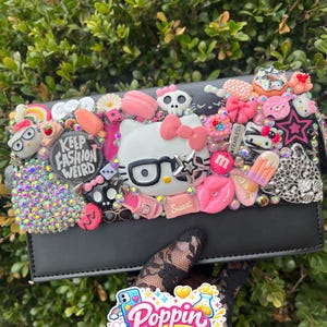 May include: Black clutch decorated with various charms, including a Hello Kitty face, a skull, and a rainbow. The clutch has the text "Keep Fashion Weird" and "Sweet". The image also shows a "Poppin Case Lab" sticker.