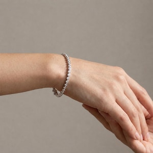 May include: A silver-toned tennis bracelet featuring a continuous row of round, clear, bezel-set stones. The bracelet encircles the wrist, showcasing a uniform design with each stone securely held in a polished metal setting, creating a classic look.
