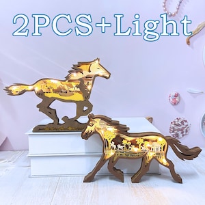 Op de afbeelding: Twee houten paarden silhouetten met een verlicht ontwerp. De paarden rennen en hebben een gedetailleerd tafereel binnen de silhouet. De tekst "2PCS+Light" staat bovenaan de afbeelding.