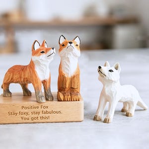 May include: Three carved wooden fox ornaments. Two are orange and white, one is white. The orange foxes stand on a wooden base with the text "Positive Fox. Stay foxy, stay fabulous. You got this!" The foxes are looking upwards.