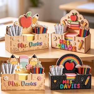 Puede incluir: Cuatro organizadores de escritorio de madera con recortes coloridos de manzanas, lápices y flores. Cada unidad tiene compartimentos llenos de marcadores y lápices de colores, una ranura central para notas adhesivas y texto personalizado como Mrs. Davies y Mrs. Chloe.
