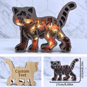 Op de afbeelding: Een houten katvormig nachtlampje met ingewikkelde uitsparingen en warme LED-verlichting. De kat is 17 cm lang en 13,3 cm hoog. Het ontwerp bevat een gedetailleerd stadsbeeld zichtbaar door de uitsparingen.