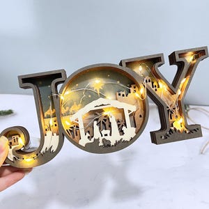 以下が含まれることがあります： 「JOY」と書かれた木製のクリスマスデコレーション。文字の「O」の中には降誕シーンが描かれています。文字は茶色で、温かみのある白色LEDライトが使用されています。降誕シーンには、星、厩、人や動物のシルエットが含まれています。