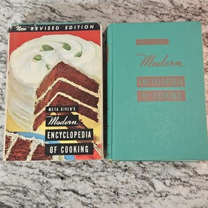 May include: Two copies of Meta Given's "Modern Encyclopedia of Cooking" cookbook. The newer edition has a picture of a chocolate cake with white frosting on the cover. The older edition has a teal blue cover with the title in gold lettering.