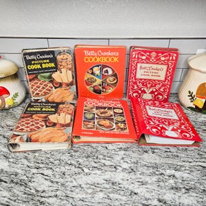 May include: A selection of vintage Betty Crocker cookbooks. The books have red, orange, and black covers, with images of food. Titles include "Betty Crocker's Picture Cook Book" and "Betty Crocker's Cookbook". The books are in various sizes.
