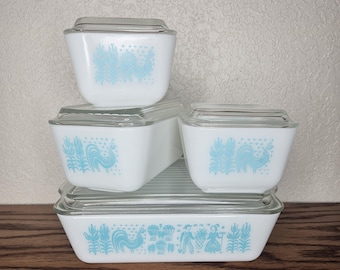 Puede incluir: Un juego de recipientes de almacenamiento de alimentos Pyrex blancos vintage con tapas de vidrio transparente. Cada recipiente presenta un diseño de gallo y floral azul claro. El juego incluye varios tamaños, apilados sobre una superficie de madera.