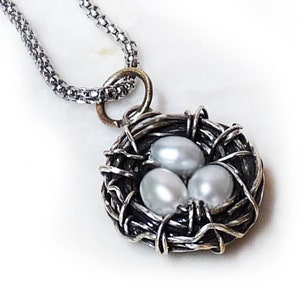 Könnte beinhalten: Eine silberne Halskette mit einem Anhänger in Form eines Vogelnests mit drei weißen Perlen darin.