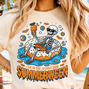 May include: A cream-coloured t-shirt featuring a skeleton wearing sunglasses, relaxing on a donut in water, holding ice cream. The shirt displays the text "SUMMERWEEN" and "FUN IN THE SPOOKY SUN!" A Halloween and summer beach theme.