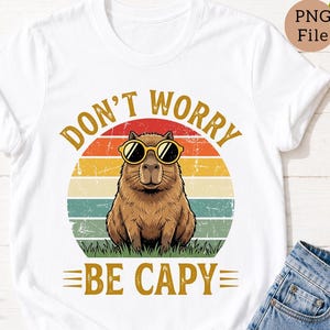 May include: White t-shirt featuring a brown capybara wearing sunglasses, set against a retro sunset design. The shirt includes the text "DON'T WORRY BE CAPY" in a vintage font. The capybara is sitting on green grass.