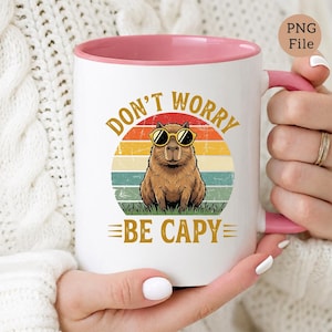 May include: White ceramic mug with a pink interior and handle. The mug features a capybara wearing sunglasses, set against a retro sunset design. Text on the mug reads "DON'T WORRY BE CAPY".