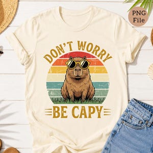 May include: Cream-colored t-shirt featuring a retro design with a capybara wearing sunglasses. The text "DON'T WORRY BE CAPY" is printed above and below the capybara illustration, set against a sunset-style background.