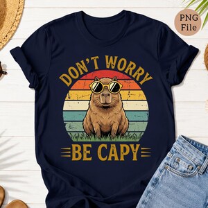 May include: Navy blue t-shirt with a graphic of a capybara wearing sunglasses, set against a sunset design. The text reads "DON'T WORRY BE CAPY" in yellow. The shirt is a casual wear item.