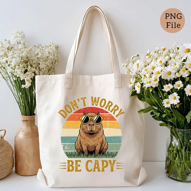May include: Cream-colored tote bag featuring a capybara wearing sunglasses, set against a retro sunset design. The text "DON'T WORRY BE CAPY" is printed above and below the capybara illustration. The bag has two handles.