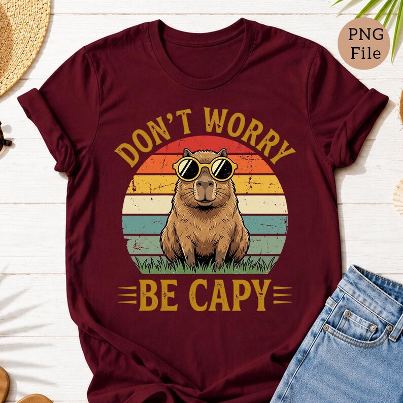 May include: Burgundy t-shirt featuring a capybara wearing sunglasses, set against a retro sunset design. The text "DON'T WORRY BE CAPY" is printed in a vintage style. The shirt is made of a soft material.