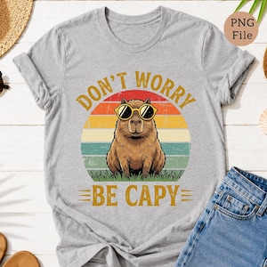 May include: A light gray t-shirt featuring a capybara wearing sunglasses, set against a retro sunset design. The text "DON'T WORRY BE CAPY" is printed in gold. The shirt is paired with blue denim jeans.