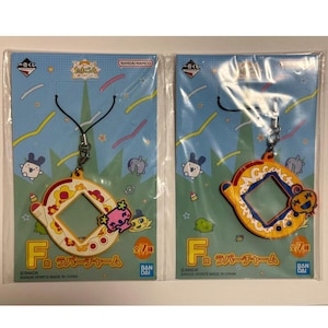 May include: Two colorful rubber charms featuring a yellow and orange Tamagotchi-like device with a blue screen. The charms are attached to black strings and are packaged on a white card with the text "F ラバーチャーム 全7種" and "BANDAI SPIRITS MADE IN CHINA".