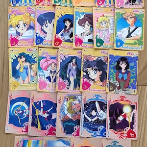 以下が含まれることがあります： 様々なキャラクターが描かれた、鮮やかな色のセーラームーンのトレーディングカードのコレクション。各カードには、キャラクターの異なるイラスト、日本語のテキスト、そして「セーラームーン」のロゴが表示されています。